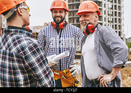 Great work! Group of builders in protective wear and red helmets are discussing new project and smiling while standing at construction site. Teamwork. Stock Photo