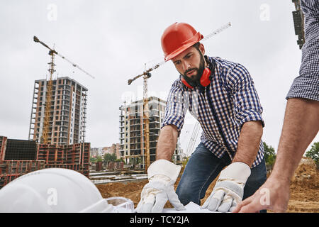 Discussing new project. Young bearded builder in red helmet looking at construction drawing and discussing it with co-worker while standing at constru Stock Photo