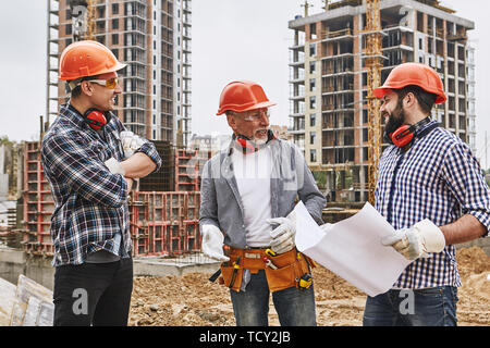 Professional team. Group of builders in protective wear and red helmets are discussing new project while standing at construction site. Teamwork. Arch Stock Photo