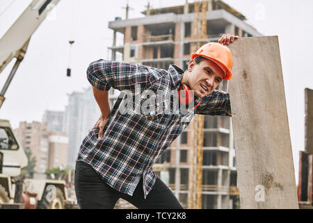 Hard work. Construction worker in protective helmet feeling back pain while working at construction site. Building construction. Pain concept. Dangero Stock Photo