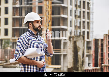 Working process. Professional young at construction builder in white protective helmet talking to crane operator by walkie talkie while standing at co Stock Photo