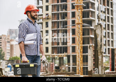 Work day is starting. Cheerful young builder in red helmet holding toolbox, protectives gloves and engineering drawings while standing at construction Stock Photo
