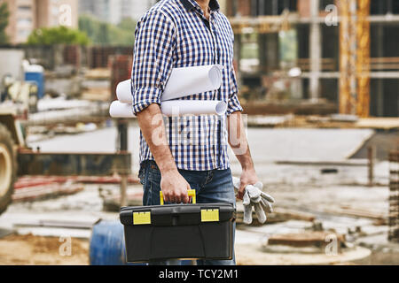 Everything what i need for work. Cropped image of a builder holding toolbox, protectives gloves and engineering drawings while standing at constructio Stock Photo