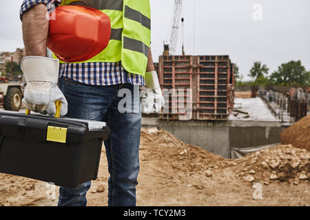 Ready to work! Cropped image of a builder in working uniform holding red helmet and carrying toolbox while standing at construction site. Building con Stock Photo