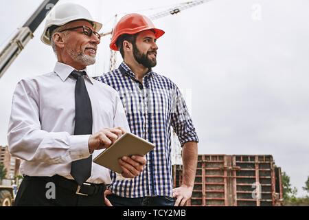 Checking work. Senior building inspector in formal wear and white helmet is holding a digital tablet and looking with young builder what is already do Stock Photo
