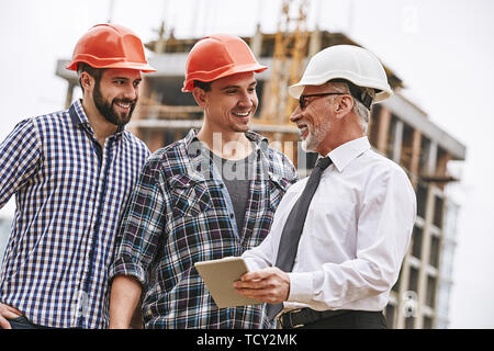 Listen carefully. Senior engineer in white helmet and formal wear is holding digital tablet and explaining building plan to young builders while stand Stock Photo