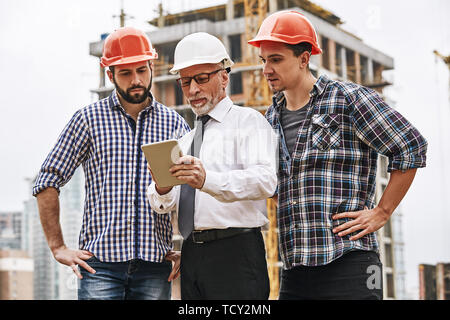 Using modern technologies. Senior engineer is looking at project plan on digital tablet with two young builders while working at construction site. Te Stock Photo