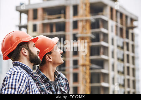 Teamwork. Two young and cheerful builders in red helmets are looking up and working at construction site. Construction concept. Building concept. Stock Photo