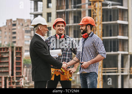 Good job! Cheerful elderly engineer in formal wear and white helmet holding engineering drawings and shaking hands of two happy young builders while s Stock Photo
