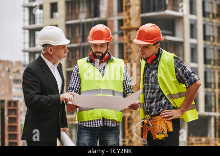 Team meeting. Main engineer in formal wear and white helmet with two young builders are discussing a construction drawing and main concept of building Stock Photo