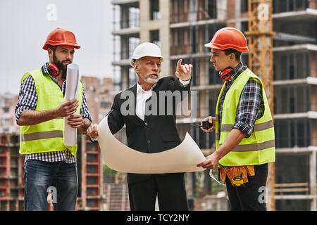 Plan of building. Main engineer in formal wear and white helmet holding a sketch of new project and discussing with two young builders the problems of Stock Photo