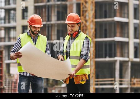 Look here! Two young builders in working uniform and helmets are holding a construction drawing in their hands and discussing it with each other while Stock Photo