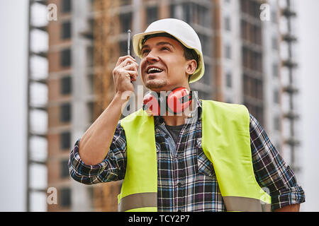 Turn left! Cheerful young builder in working uniform, yellow protective helmet talking to crane driver by walkie-talkie, looking up and smiling while  Stock Photo