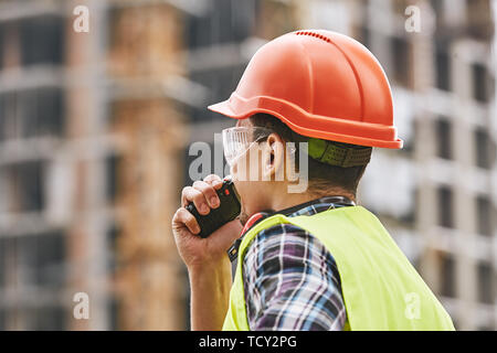 Giving instructions. Cropped photo of builder in working uniform and red protective helmet talking to crane operator by walkie talkie while standing a Stock Photo