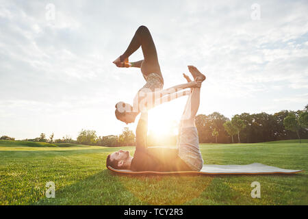Balancing Yoga Poses. Healthy and young couple practicing acro yoga together in the morning park. Man lying on a mat and balancing woman on extended a Stock Photo