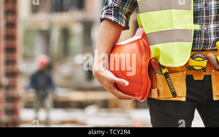 Construction worker. Cropped photo of male professional builder in working uniform with construction tools holding a safety red helmet while standing  Stock Photo
