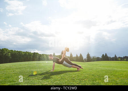 Practicing acro yoga together. Strong man is staying in plank on a grass in nature and woman is doing yoga asana by leaning on his back. Sunny morning Stock Photo