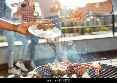 Perfect day for barbecue. Cropped photo of male hand putting hot barbecued meat on the plate while standing on the roof with friends. Barbecue concept Stock Photo
