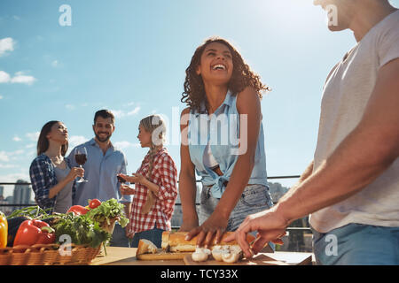 Summer picnic. Two young and cheerful friends in casual clothes preparing food for barbeque party and smiling while standing on the roof. Having fun.  Stock Photo