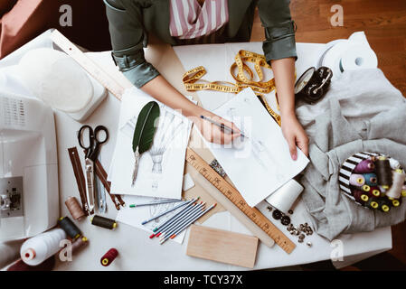Taking new ideas into paper . Top view of fabulous young woman tailor with sewing machine is thinking about new dress. Tailor items are at the table.  Stock Photo