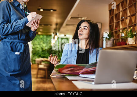 Stylish mixed race office worker in blue suit is sitting at the table. She choose what she wants to eat and makes an order while the waiter taking it Stock Photo