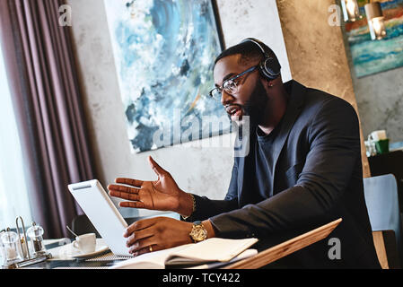 Serious stylish young Afro American businessman in dark-blue jacket and glasses sitting at the table, indoors, wearing wireless earbuds, chatting with Stock Photo