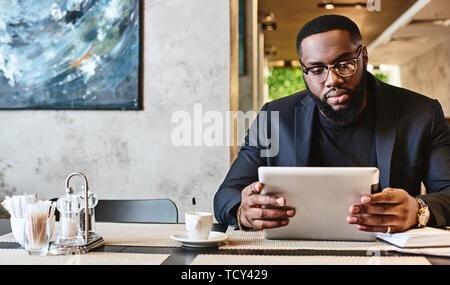 Portrait of focused African-American office manager in dark-blue jacket and glasses, sitting at cafe table with tablet pc, reading important documents Stock Photo