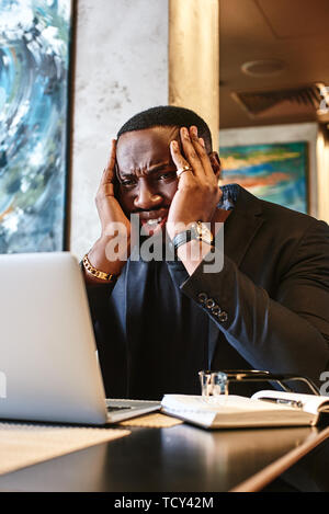 Focused African-American office manager in dark-blue jacket sitting at cafe table with laptop, reading important documents with puzzled expression and Stock Photo