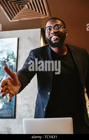 Portrait of dark-skinned businessman in dark-blue jacket and glasses standing, welcoming his business partner indoors, smiling, offering handshake. Yo