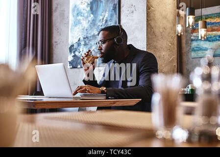 Portrait of dark-skinned businessman in dark-blue jacket and glasses sitting at the table, indoors, wearing headphones, drinking coffee, working with  Stock Photo