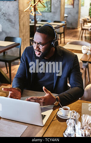 Portrait of dark-skinned businessman in dark-blue jacket and glasses sitting at the table, indoors, wearing headphones, working with his laptop comput Stock Photo
