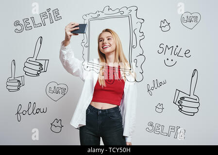 I love selfie! Cute young woman with blond hair making selfie by her smartphone and smilingwhile standing against grey background with different hand  Stock Photo