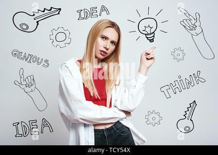 I have an idea! Pretty blonde woman in casual clothing pointing pointing at lightbulb while standing against grey background with hand drawn doodles o Stock Photo
