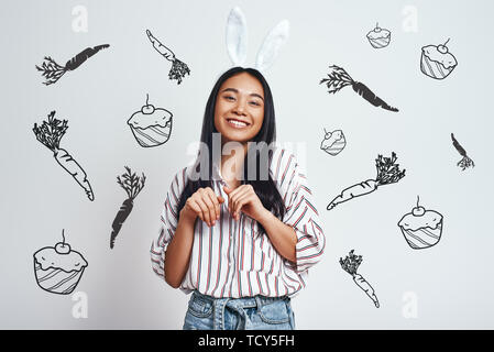 Cuter than any bunny. Attrative young asian woman in bunny ears is smiling while standing against grey background with hand drawn carrots and cupcakes Stock Photo