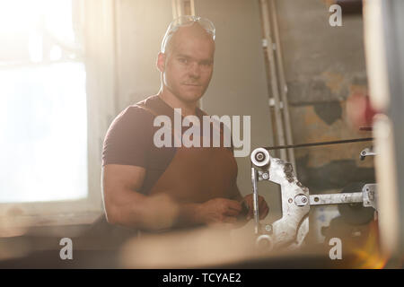 Portrait of serious confident master of knife making with dirty face keeping safety goggles on bald head standing by grinding machine Stock Photo