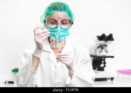 Young attractive concentrated female scientist in protective eyeglasses, mask and gloves dropping a red liquid substance into the test tube with a pipette in the laboratory . Stock Photo