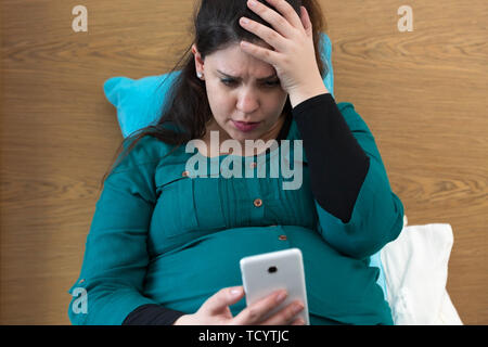Frustrated pregnant woman sitting on bed using her smartphone Stock Photo