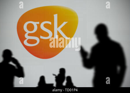 The GSK logo is seen on an LED screen in the background while a silhouetted person uses a smartphone in the foreground (Editorial use only) Stock Photo