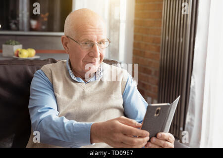 Senior man using a smartphone and smiling while sitting on couch at home. Stock Photo