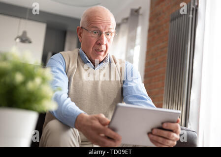 Senior man surprised using a digital tablet while sitting on couch at home Stock Photo