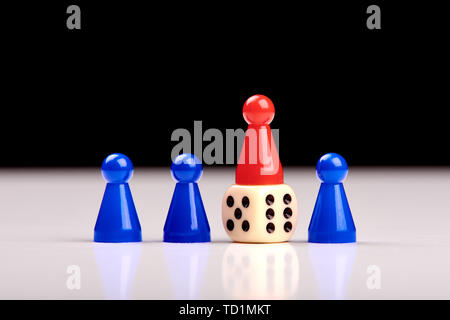 Three blue game pieces and between them one red piece stands on a dice as a winner or leader. Blurred black and white background Stock Photo
