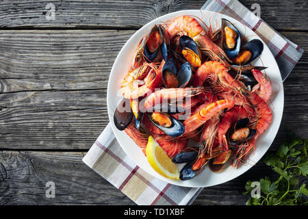 mix seafood - mussels, clams and king prawns on a white plate on a rustic wooden table, horizontal view from above, close-up