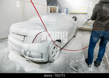 Male worker wash the car with high pressure washer. Car wash with foam in car wash station. A man sprays foam on car from a special spray. Stock Photo