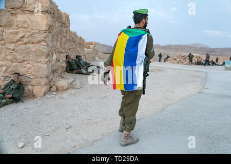 Masada, Israel. 23 October 2018: Soldier walking with a descending green, red, yellow, blue and whit stripes flag of Druze during the war games. Stock Photo