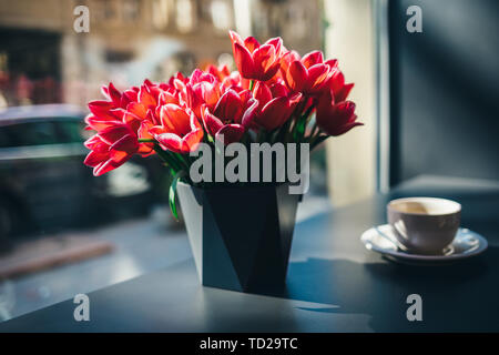 Bouquet of beautiful red tulip flowers in vase on table by the window next to cup of coffee, sun shines through glass. Stock Photo