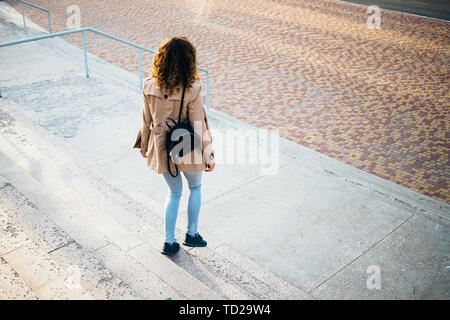 Stylish young woman dressed in beige coat with black backpack goes down the stairs outdoors, rear view. Stock Photo