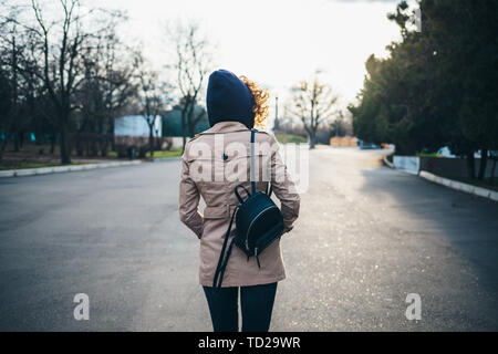 Rear view young woman with curly hair wearing beige trench coat hoodie and backpack walking on road in city. Stock Photo