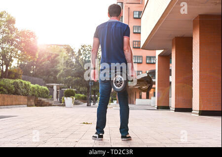 A man in jeans and sneakers on a hoverboard ride in the city. Happy boy riding around at sunset. Modern electronics for relaxation and entertainment Stock Photo