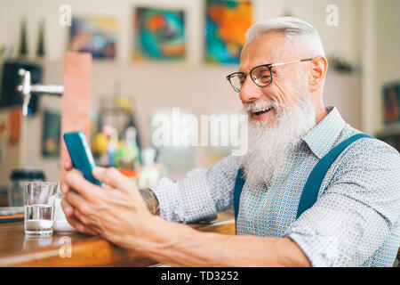 Hipster man using mobile phone while sitting in a bar - Senior male watching on new trends smartphone apps for social media Stock Photo