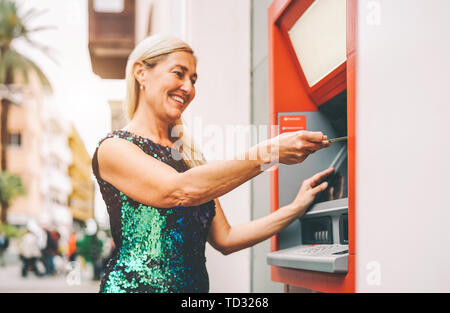 Happy mature woman withdraw money from bank cash machine with debit card - Senior female doing payment with credit card in ATM Stock Photo